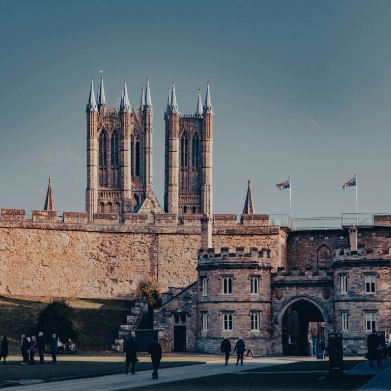 Historic castle with tall towers, stone walls, and flags flying, set against a blue sky.